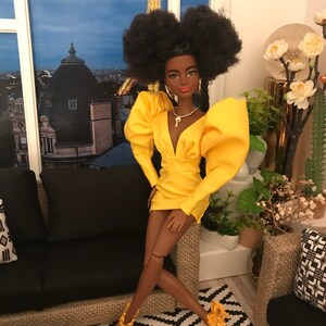 May include: A fashion doll with dark brown skin, black afro hair, and a yellow dress with puffed sleeves. The doll is wearing gold jewellery and gold heels. The doll is sitting on a black sofa with a patterned rug in the background.