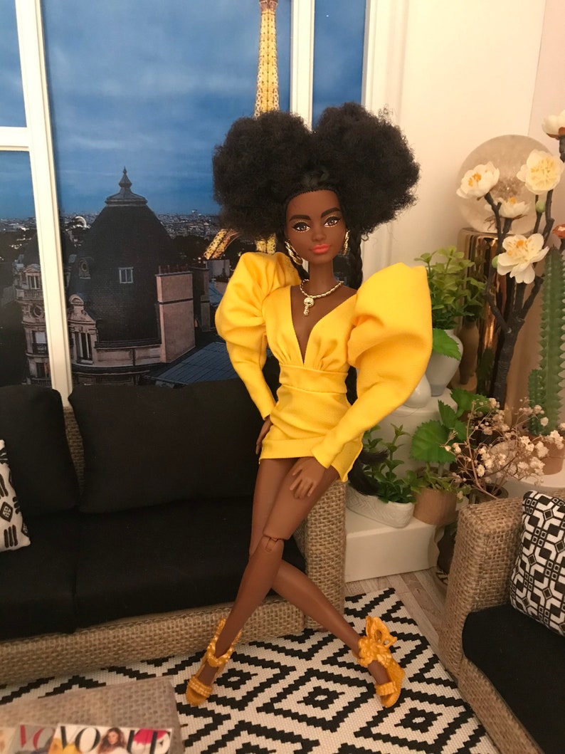 May include: A fashion doll with dark brown skin, black afro hair, and a yellow dress with puffed sleeves. The doll is wearing gold jewellery and gold heels. The doll is sitting on a black sofa with a patterned rug in the background.