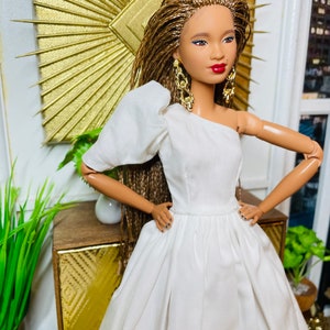 May include: A doll with brown braided hair wearing a white one-shoulder dress and gold earrings. The doll is standing in front of a gold sunburst mirror and a wooden cabinet.