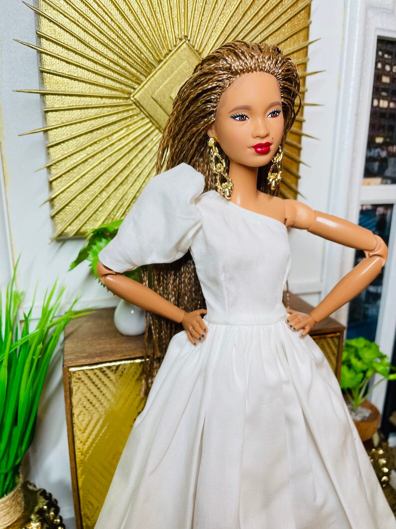 May include: A doll with brown braided hair wearing a white one-shoulder dress and gold earrings. The doll is standing in front of a gold sunburst mirror and a wooden cabinet.
