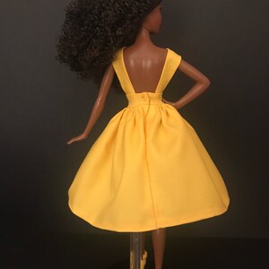May include: A yellow sleeveless dress with a low back and a gathered skirt. The dress is on a doll with brown curly hair.