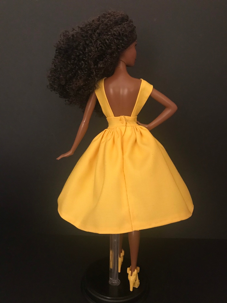 May include: A yellow sleeveless dress with a low back and a gathered skirt. The dress is on a doll with brown curly hair.