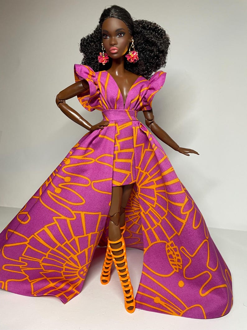 May include: A fashion doll wearing a purple and orange dress with a high slit and matching orange high-heeled boots. The doll has dark skin, black curly hair, and pink earrings.