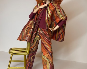 May include: A redheaded doll wearing a maroon top and pants with a gold and orange swirl pattern. The doll is wearing a matching cape with the same pattern. The doll is standing next to a gold chair.
