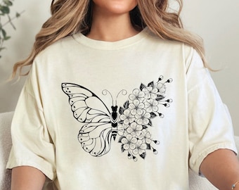 May include: A white t-shirt with a black and white graphic of a butterfly with flowers. The butterfly is in the center of the shirt and the flowers are on the right side of the butterfly.