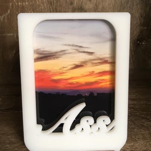 May include: White plastic picture frame with a sunset photo inside. The frame has a cutout of the word "Ass" in the bottom right corner.
