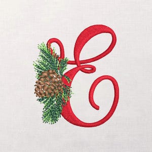 May include: Embroidered red letter "E" with a festive pinecone and evergreen sprig design. The letter is in a cursive font, and the pinecone is brown with green accents. The background is a light, textured fabric.