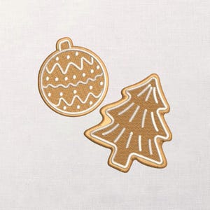 May include: Two embroidered Christmas decorations: a round bauble and a Christmas tree. The ornaments are tan with white and gold detailing. The bauble has a zig-zag and dot pattern. The tree has vertical lines.