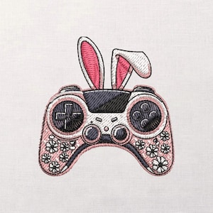 Könnte beinhalten: Gestickter rosa-schwarzer Videospiel-Controller mit weißen floralen Akzenten. Der Controller hat ein kreuzförmiges Steuerkreuz, Tasten und Hasenohren. Das Design befindet sich auf weißem Hintergrund.