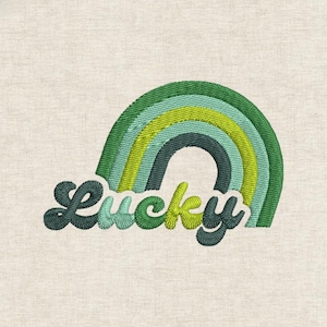 Puede incluir: Diseño bordado con un arcoíris verde y azul con la palabra "Lucky" en una fuente cursiva. El diseño está cosido sobre una tela beige claro, creando una apariencia texturizada. El arcoíris tiene múltiples capas de verde y azul.