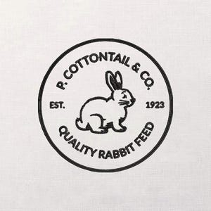 Puede incluir: Un logotipo bordado en blanco y negro con un conejo dentro de un marco circular. El texto dice "R. Cottontail & Co. Est. 1923 Quality Rabbit Feed". El logotipo está sobre un fondo de tela blanca.