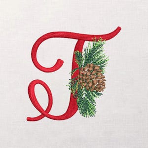 Peut inclure: Lettre "J" rouge brodée avec une pomme de pin décorative et des brins de conifères. Le motif présente un aspect détaillé et texturé, idéal pour les créations sur le thème des fêtes ou les cadeaux personnalisés. La lettre est en cursive.