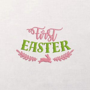 May include: White fabric with embroidered text and design. The words "First Easter" are in pink and green, with a pink bunny and floral accents. The embroidery is detailed and the design is centered.