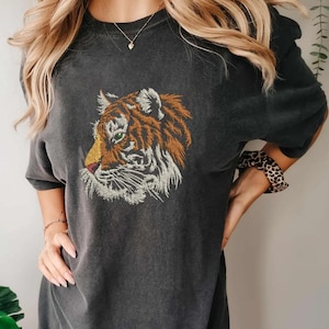 May include: Dark gray t-shirt featuring an embroidered tiger head. The tiger design showcases orange, brown, and white details with green eyes. The shirt has short sleeves and a relaxed fit, suitable for casual wear.