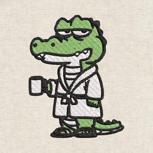 Op de afbeelding: Een geborduurde patch met een groen krokodil die een witte badjas draagt en een kleine witte mok vasthoudt. De krokodil heeft een ontspannen uitdrukking met halfgesloten ogen, omlijnd met dikke zwarte draad op een lichte stoffen achtergrond.