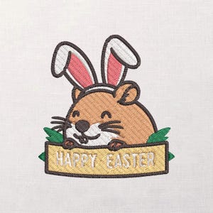 May include: Embroidered design of a brown hamster wearing bunny ears, holding a sign that reads "HAPPY EASTER". The hamster has a smiling face and black whiskers. Green leaves are on either side of the sign. The design is on a white background.