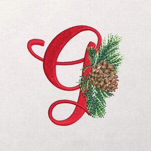 Puede incluir: Letra "G" roja bordada con un diseño decorativo de piña y rama de hoja perenne. La letra está en cursiva, y la piña es marrón con acentos verdes y amarillos. El fondo es una tela clara y texturizada.