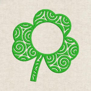 Puede incluir: Un diseño de trébol verde con una abertura circular en el centro. El trébol está relleno de patrones decorativos en espiral. El diseño está bordado sobre una tela beige claro, ideal para manualidades y decoración del Día de San Patricio.