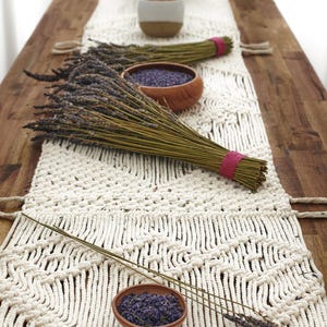 Peut inclure: Chemin de table en macramé crème avec des bouquets de lavande séchée liés par un ruban rose. De petits bols en bois contiennent des boutons de lavande en vrac. La scène est sur une table en bois.