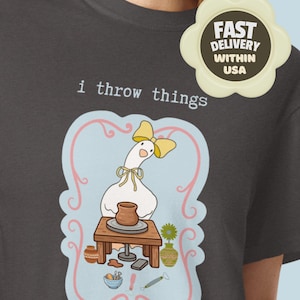 May include: A dark grey t-shirt featuring a graphic of a white goose wearing a yellow bow, sitting at a wooden pottery wheel with a clay pot. The text "i throw things" is printed above the goose, surrounded by a light blue frame with pink swirls.