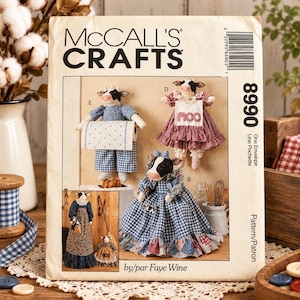 Op de afbeelding: Een McCall's Crafts 8990 naaipatroon-envelop met illustraties van drie handgemaakte koeienpoppen in diverse outfits. De poppen dragen jurken met blauwe vichy-ruitjes, bloemen en patronen, waarbij één pop een bordje vasthoudt met de tekst moo.