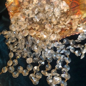 May include: A decorative, iridescent, gold-toned bowl overflowing with strands of clear, faceted crystal beads. The crystals are strung together, creating a cascading effect. The bowl sits on a dark green velvet surface.