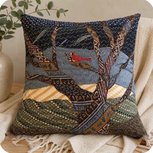 May include: A square throw pillow featuring a patchwork design of a landscape with a red cardinal bird perched on a tree branch. The scene includes various fabric patterns in shades of navy blue, brown, gold, and green with intricate stitching.