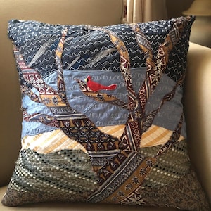 May include: A decorative square pillow with a quilted design featuring a tree and a red cardinal. The pillow has a patchwork design with various fabric patterns in shades of blue, brown, and yellow. The cardinal is perched on a branch.