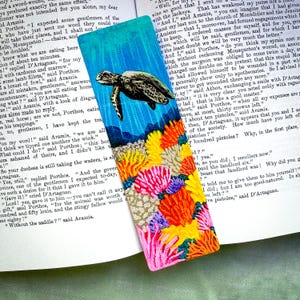 May include: Hand-painted bookmark featuring a sea turtle swimming above colorful coral. The bookmark has a blue background and is rectangular. The coral is in shades of red, orange, yellow, and pink.