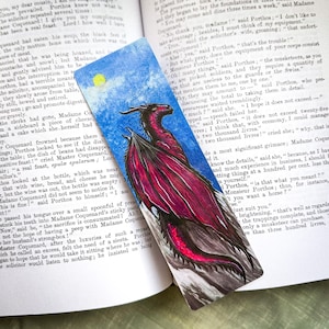 May include: A red dragon bookmark with black wings and a blue background. The dragon is flying through the air with its mouth open and its claws extended. The bookmark is made of a sturdy material and is perfect for any book lover.