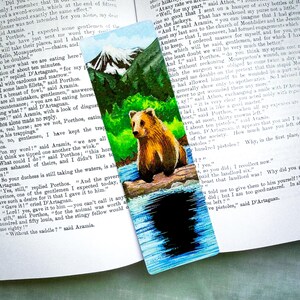 May include: Hand-painted bookmark featuring a brown bear in a forest scene with a mountain and a river. The bear is the focal point, set against a backdrop of green trees and a blue sky. The bookmark is rectangular.