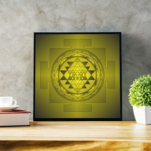 May include: A framed Sri Yantra print in yellow and black tones. The artwork is displayed on a wooden surface, alongside a book, a teacup, and a small potted plant. The background is a textured grey wall. The print is approximately 30 cm square.