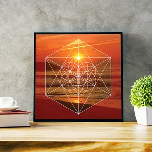 May include: Framed art print featuring a geometric design overlaid on a sunset beach scene. The artwork is in a black frame and rests on a wooden surface. A white teacup and saucer sit on a red book, with a small potted plant nearby.