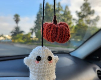 May include: A crocheted white ghost ornament with black bead eyes and pink stitched cheeks, paired with a small crocheted orange pumpkin, both hanging from a black string designed to be suspended from a vehicle rearview mirror.