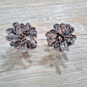 May include: Two decorative pine cone shaped objects with a white and gray marbled design. The objects are on a light wood surface.