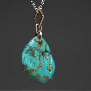 May include: A turquoise gemstone pendant necklace. The pendant is a polished, irregular shape with brown veining. It hangs from a silver chain with a geometric gold-colored connector. The background is a solid, dark gray.