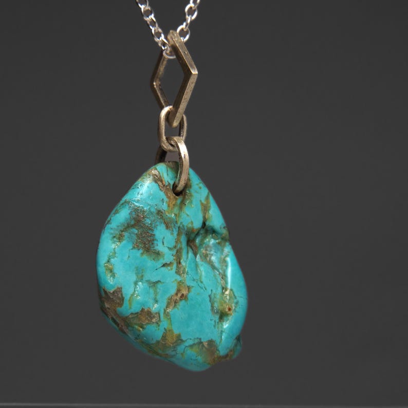 May include: A turquoise gemstone pendant necklace. The pendant is a polished, irregular shape with brown veining. It hangs from a silver chain with a geometric gold-colored connector. The background is a solid, dark gray.
