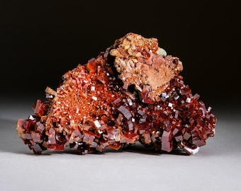 May include: A cluster of red vanadinite crystals with a rough, earthy texture. The crystals are sharp and angular, with a deep red color. The crystals are arranged in a cluster, with some crystals overlapping others.