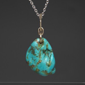 May include: A turquoise pendant necklace with a silver chain. The pendant is a freeform, polished stone with a vibrant blue-green color and brown matrix patterns. The chain is delicate and hangs from a silver bail.