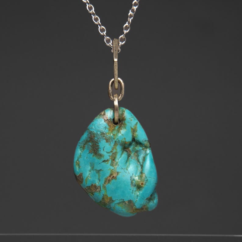 May include: A turquoise pendant necklace with a silver chain. The pendant is a freeform, polished stone with a vibrant blue-green color and brown matrix patterns. The chain is delicate and hangs from a silver bail.