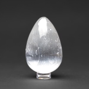 Op de afbeelding: Een helder, glad, eivormig selenietkristal op een heldere glazen voet. Het kristal heeft een licht troebele uitstraling met een subtiele glans.