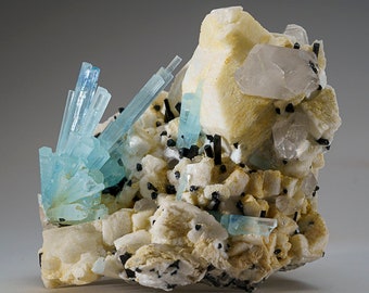 May include: A cluster of aquamarine crystals with a light blue color, set in a matrix of white and tan rock. The crystals are sharp and well-defined, and some are translucent. There are also black crystals scattered throughout the matrix.