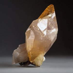 May include: A large, clear, orange-brown crystal with a pointed tip. The crystal is sitting on a white surface.