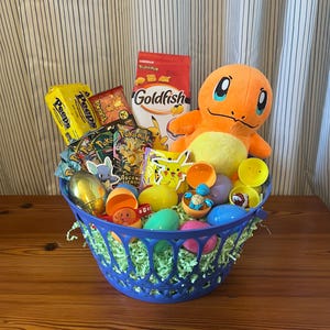 May include: A blue Easter basket filled with treats and toys. The basket contains a plush Charmander, a bag of Goldfish crackers, Peeps, and various Pokemon-themed items. Colorful plastic eggs are scattered throughout the basket.