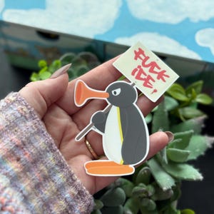 Puede incluir: Una pegatina con un pingüino de dibujos animados sosteniendo un cartel que dice "FUCK ICE!". El pingüino es gris, negro y amarillo, con un pico y patas naranjas. La pegatina se sostiene en una mano.