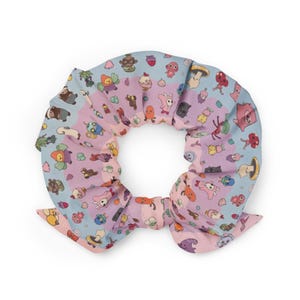 May include: A colorful scrunchie with a whimsical pattern of cartoon characters in shades of blue, pink, and purple. The fabric is gathered and tied into a bow, creating a playful accessory for hair styling.