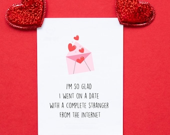 Könnte beinhalten: Eine weiße Grußkarte mit dem Text "I'M SO GLAD I WENT ON A DATE WITH A COMPLETE STRANGER FROM THE INTERNET" und einer rosa Umschlagillustration mit Herzen. Zwei rote Glitzerherzen auf rotem Hintergrund.