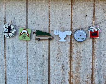 May include: A string of Christmas ornaments with various designs, including a cat with lights, a hat with the text "You Serious Clark?", a car with a Christmas tree on top, a moose, a gas gauge that reads "Shitter", and a jar of jelly.