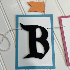 May include: A white banner with a blue border and a black letter "B" in a gothic font. The banner is strung with white twine and has a small orange flag with a silver pole.