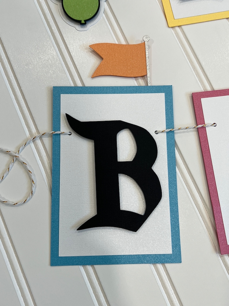 May include: A white banner with a blue border and a black letter "B" in a gothic font. The banner is strung with white twine and has a small orange flag with a silver pole.